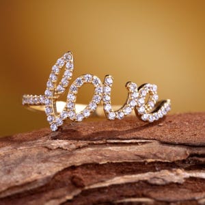 May include: A gold-colored ring spelling out the word "love" in cursive script. The letters are encrusted with small, clear gemstones. The ring is set against a warm, golden background and rests on a piece of wood.