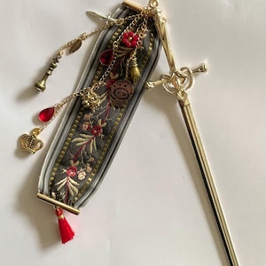 May include: A gold-colored sword-shaped hair stick with a decorative ribbon. The ribbon is gray with a floral pattern and gold trim, adorned with charms including a crown, teardrops, and a chess piece. A red tassel hangs from the bottom.