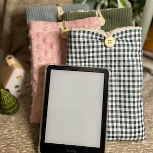 Peut inclure: Un lecteur Kindle dans un étui transparent est présenté devant plusieurs étuis en tissu. Les étuis sont de différentes couleurs et textures, dont rose à pois, vert en velours côtelé et vichy vert et blanc. Chaque étui a une fermeture à bouton.