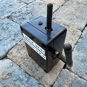 May include: A black rectangular device with a vertical antenna and a smaller angled antenna. The device has the text "MCZ Wifi Modul V2.0" on a white label. The device is on a grey stone surface.