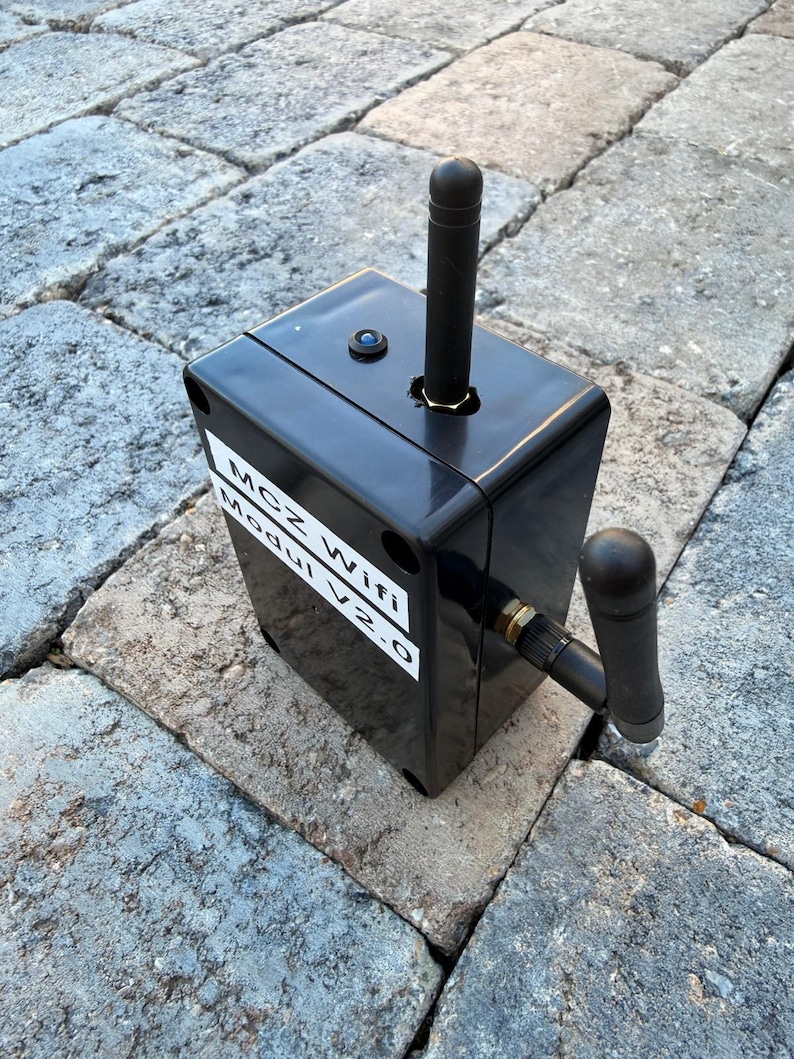 May include: A black rectangular device with a vertical antenna and a smaller angled antenna. The device has the text "MCZ Wifi Modul V2.0" on a white label. The device is on a grey stone surface.