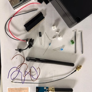 Könnte beinhalten: Eine Ansammlung elektronischer Komponenten, darunter eine Leiterplatte, Drähte, eine schwarze Box und verschiedene Anschlüsse. Eine schwarze Antenne und eine kleine Leiterplatte mit einem goldenen Anschluss sind ebenfalls vorhanden. Die Komponenten sind auf einer weißen Oberfläche angeordnet.