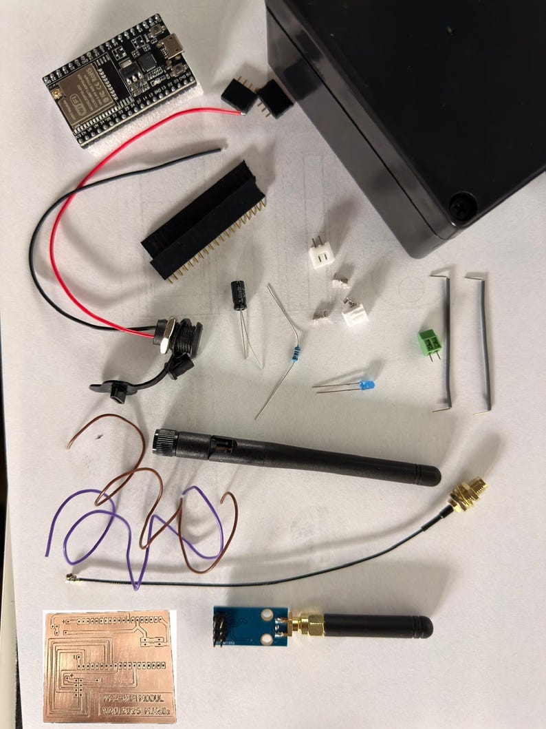 May include: A collection of electronic components, including a circuit board, wires, a black box, and various connectors. A black antenna and a small circuit board with a gold connector are also present. The components are arranged on a white surface.