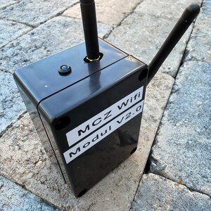 May include: A black rectangular device with two black antennas and a small blue light. The device has a white label with the text "MCZ Wifi Modul V2.0". The device is sitting on a stone surface.