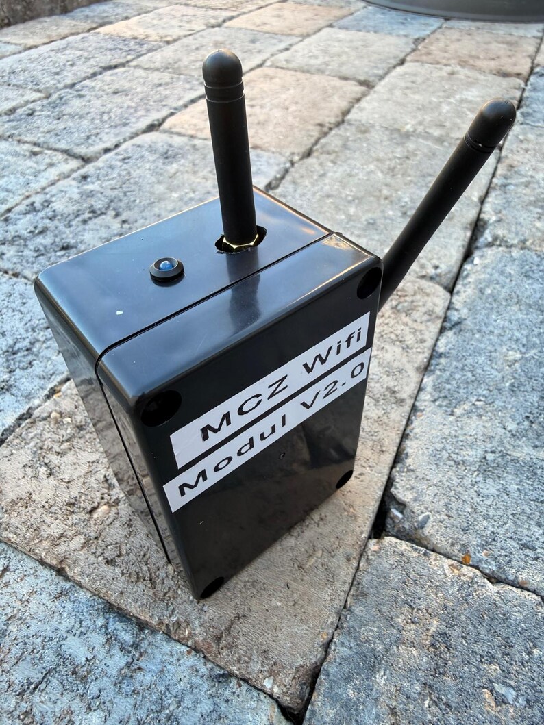 May include: A black rectangular device with two black antennas and a small blue light. The device has a white label with the text "MCZ Wifi Modul V2.0". The device is sitting on a stone surface.