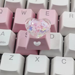 May include: A close-up of a keyboard with pink and white keys. A clear heart-shaped keycap filled with iridescent stars sits on a pink key. The pink key has a small white heart on it.