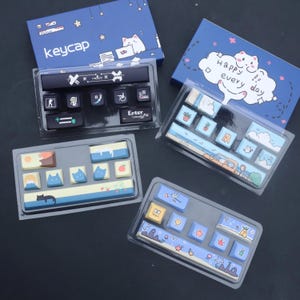 May include: A selection of custom keycaps in clear plastic packaging, with blue boxes featuring cat illustrations and text. The keycaps have various designs, including cats, landscapes, and other graphics. The word "keycap" is printed on one box.