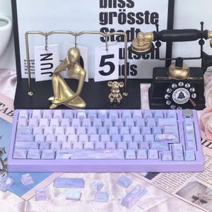 May include: A lavender mechanical keyboard with white and purple keycaps. The keyboard has a gradient design and a silver volume knob. Additional keycaps are scattered around the keyboard. Decorative items include a gold figurine and a vintage telephone.