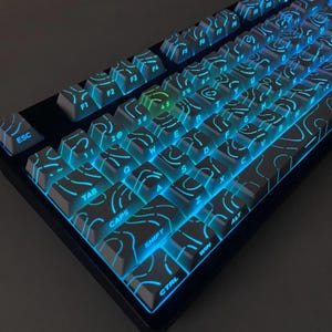 Puede incluir: Un teclado mecánico en blanco y negro con retroiluminación azul y verde. Las teclas tienen un diseño topográfico. El teclado tiene teclas etiquetadas ESC, TAB, CAPS, SHIFT, CTRL, WIN y teclas de función.