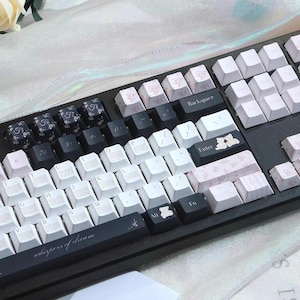 Może przedstawiać: Czarno-biała klawiatura mechaniczna z niestandardowymi nakładkami na klawisze. Nakładki na klawisze mają kwiatowe i niedźwiedzie wzory z napisem "whispers of dream". Klawiatura ma mieszankę białych i czarnych klawiszy.