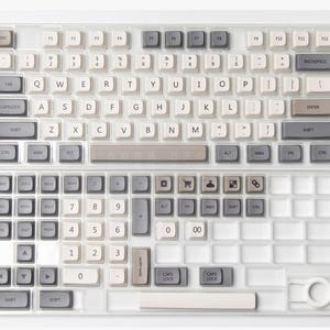 May include: A split, clear keyboard with white, gray, and beige keycaps. The keyboard has a numeric keypad and function keys. Some keys have Japanese characters. The keyboard is designed for a computer.