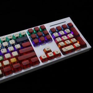 May include: A mechanical keyboard with a white frame and translucent red and white keycaps. The keys are backlit with various colours, including blue, green, and red. The keyboard has a standard layout with function keys and a number pad.