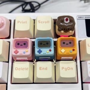 Puede incluir: Primer plano de un teclado blanco con teclas personalizadas. Las teclas incluyen un diseño de consola de juegos retro rosa, azul y amarillo, un postre de chocolate y teclas etiquetadas como "Print", "Scroll", "Delete", "End" y "PgDn".