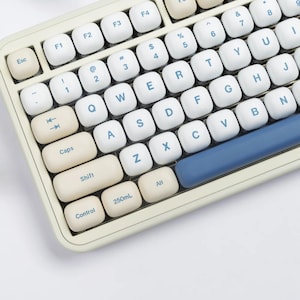 May include: Close-up of a cream-colored mechanical keyboard with white and blue keycaps. The keyboard has a retro design with rounded keys and a blue space bar. The keys are labeled with letters, numbers, and symbols.