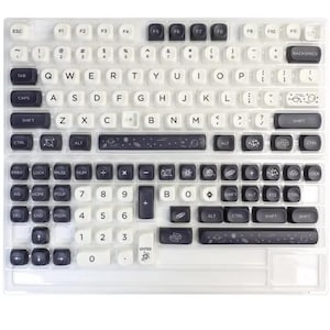 Puede incluir: Un teclado con teclas blancas y gris oscuro. Las teclas están dispuestas en un diseño QWERTY estándar. Algunas teclas tienen gráficos temáticos espaciales. El teclado está cubierto con una cubierta de plástico transparente.