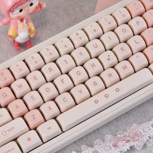 May include: A white and pink mechanical keyboard with light pink keycaps. The keyboard has a standard QWERTY layout with white lettering. A small figurine in a pink outfit is in the background.