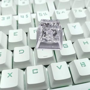 May include: A clear, custom keycap with a silver interior, hovering above a white keyboard. The keycap is transparent, allowing a view of the silver design within. The keyboard keys are white with black lettering.