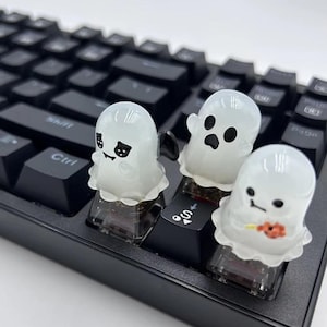 May include: Three white ghost-shaped keycaps with black facial features and a ruffled base sit on a black keyboard. One ghost has a small black wing, another holds a tiny red flower, and the third has a sad expression.