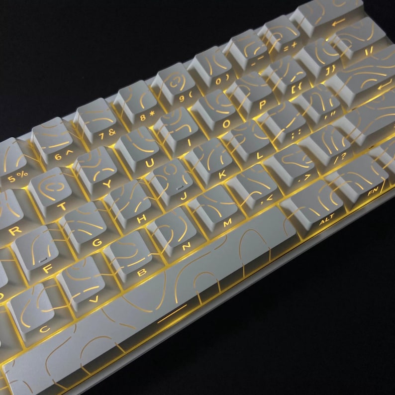 May include: A white mechanical keyboard with illuminated keys. The keycaps feature a gold topographic design. The keyboard is angled, showing the keys from the bottom left to the top right. The keys are lit with a warm, yellow light.