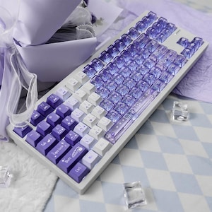 May include: A white and purple mechanical keyboard with transparent keycaps. The keyboard has a gradient design, transitioning from white to purple. Some keys have icons. The keyboard is on a light blue and white checkered surface.
