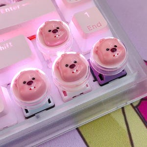 May include: Close-up of a mechanical keyboard with custom keycaps. The keycaps are shaped like pink bear faces with black eyes and a small red mouth. The keyboard keys are white and pink, with the word "Enter" visible on one key.