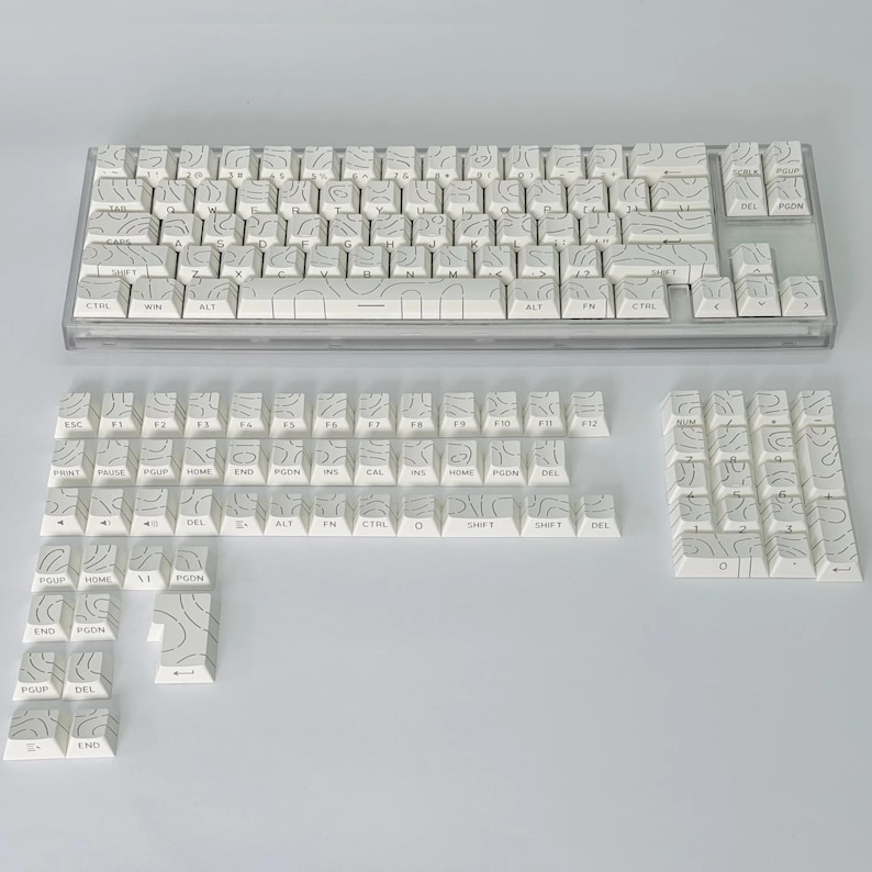 May include: A white mechanical keyboard with keycaps featuring a topographic design in grey. The keyboard is shown with extra keycaps. The keys have various labels, including letters, numbers, and function keys.