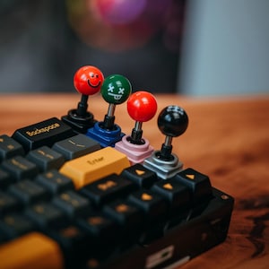 Könnte beinhalten: Nahaufnahme einer schwarzen mechanischen Tastatur mit individuellen Joystick-Tastenkappen. Die Tastenkappen sind rot, grün und schwarz mit Smiley- und X-Augen-Gesichtern. Die Eingabetaste ist gelb. Die Tastatur liegt auf einer Holzoberfläche.