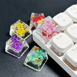 May include: A collection of clear, square-shaped keycaps with colorful dried flowers and gold flakes encased within. The keycaps are in various colors, including yellow, red, pink, purple, and teal. Some keycaps are placed on a white keyboard.