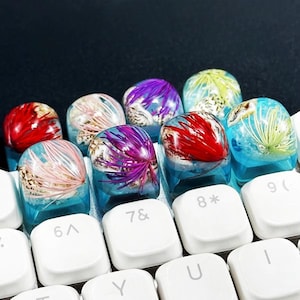May include: A set of custom keyboard keycaps with artistic designs. The keycaps feature embedded elements, including red, purple, and green floral-like shapes, set in a clear, blue resin. The keycaps are on a white keyboard. The keycaps are a unique keyboard accessory.