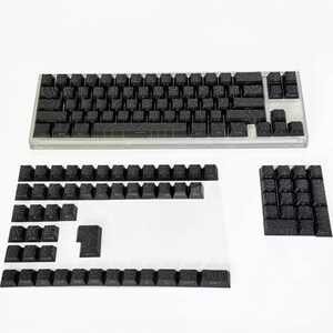 May include: A black mechanical keyboard with a clear case and a set of detached keycaps. The keycaps feature a topographic design. The keyboard is on a white surface.