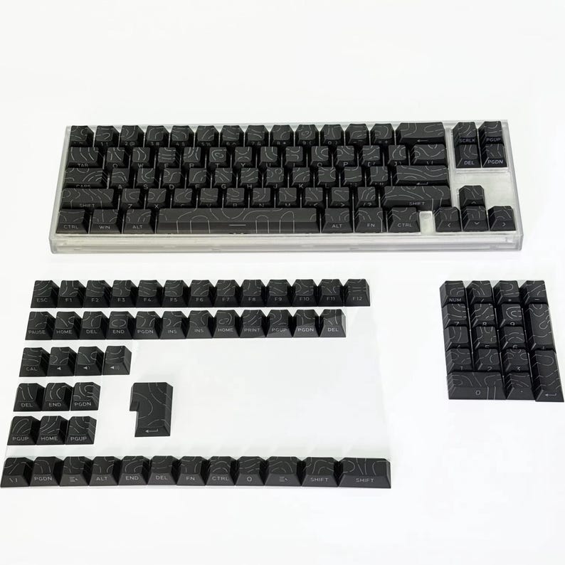 May include: A black mechanical keyboard with a clear case and a set of detached keycaps. The keycaps feature a topographic design. The keyboard is on a white surface.