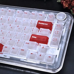 Könnte beinhalten: Eine transparente mechanische Tastatur mit weißen und roten Tastenkappen. Die Tastatur hat einen transparenten Rahmen und verfügt über eine Vielzahl von Symbolen und Zeichen auf den Tasten. Einige Tasten haben Cartoon-Bären- und Geisterdesigns. Die Tastatur hat einen Lautstärkeregler.