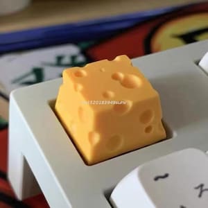 May include: A yellow cheese-shaped keycap with holes, set on a light gray keyboard. The keycap is a cube, and the keyboard keys are white and gray.
