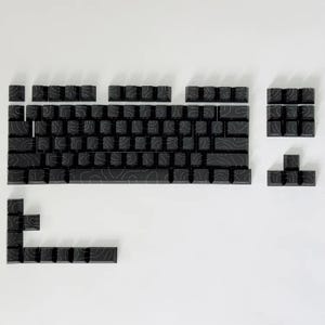 May include: Black mechanical keyboard keycaps with a topographic design. The keycaps are arranged on a white surface, showcasing a full set of keys, including the space bar, number keys, and function keys. The keycaps have a matte finish.