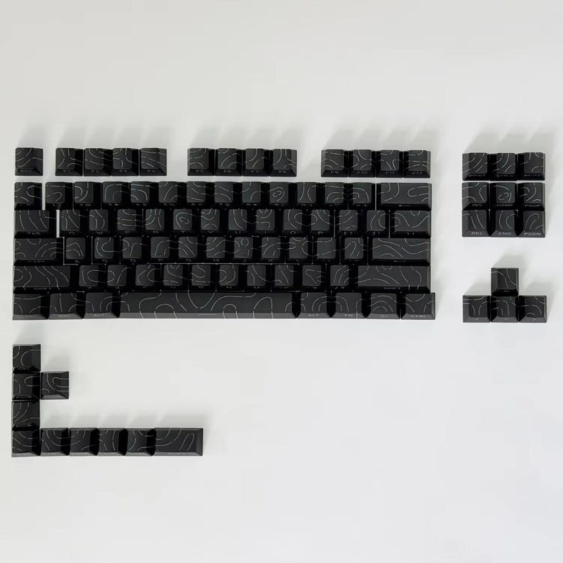 May include: Black mechanical keyboard keycaps with a topographic design. The keycaps are arranged on a white surface, showcasing a full set of keys, including the space bar, number keys, and function keys. The keycaps have a matte finish.