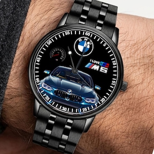 May include: Black wristwatch with a black dial featuring a blue BMW car graphic. The watch face includes the BMW logo, the text "I LOVE BMW M5", and a speedometer. The watch has a black metal band.