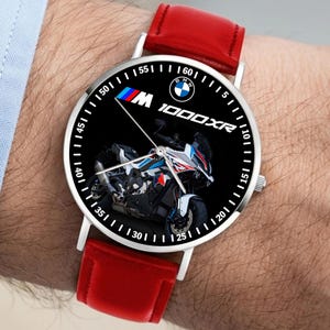 Puede incluir: Reloj de pulsera plateado con esfera negra con un gráfico de motocicleta. El reloj tiene una correa de cuero roja y el logotipo de BMW. La esfera incluye el texto "M 1000XR" y marcadores de minutos blancos.