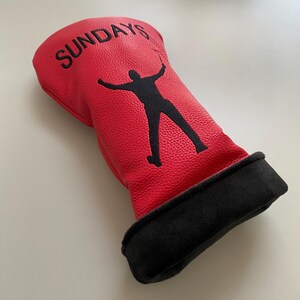 May include: Red golf club head cover with the word "SUNDAYS" in black lettering. A black silhouette of a golfer is featured on the cover. The bottom of the cover is black.
