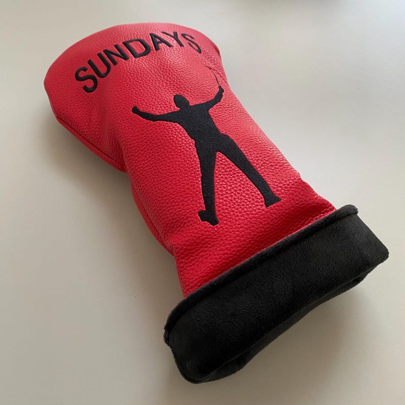 May include: Red golf club head cover with the word "SUNDAYS" in black lettering. A black silhouette of a golfer is featured on the cover. The bottom of the cover is black.