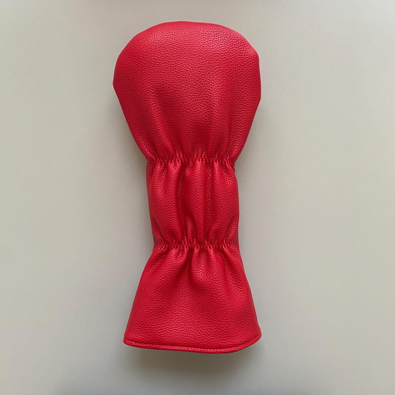 May include: A bright red golf club head cover. The cover is made of a textured material and has a rounded top and a cinched middle section. The bottom flares out slightly. The cover is designed to protect golf clubs.