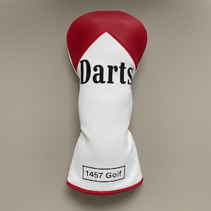 Pode incluir: Uma capa para a cabeça do taco de golfe branca e vermelha com a palavra "Darts" em letras pretas. A capa tem uma superfície texturizada e uma borda vermelha. A parte inferior da capa tem o texto "1457 Golf" num retângulo preto.