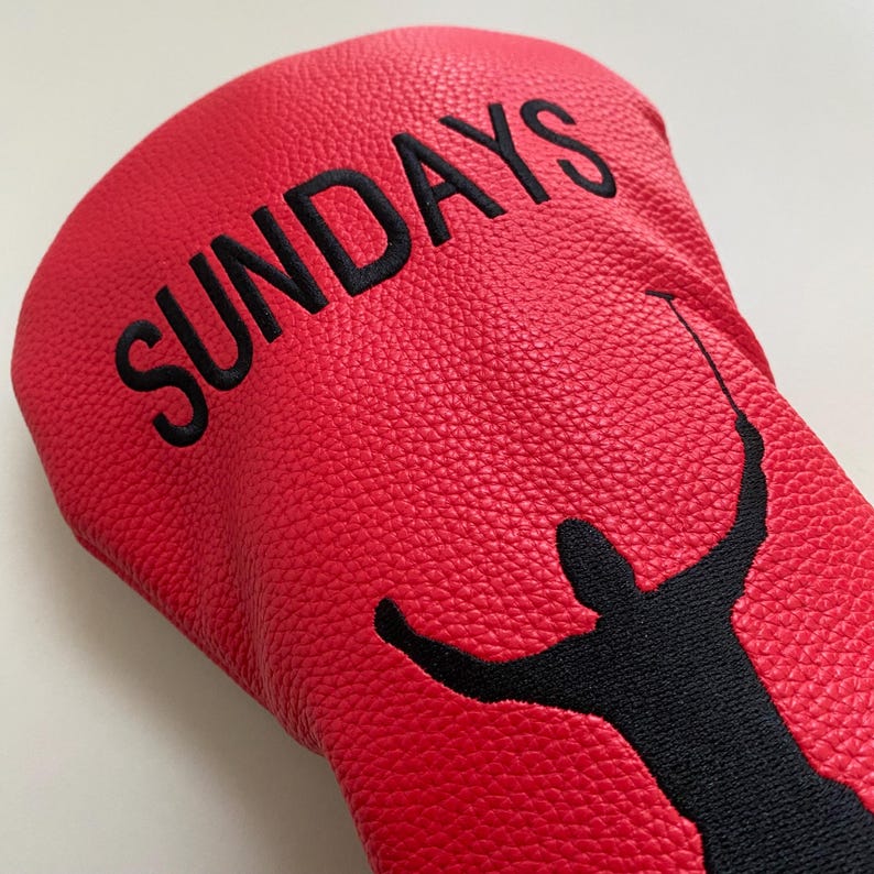 May include: Red golf club head cover with the word "SUNDAYS" embroidered in black. A black silhouette of a golfer is featured on the cover, with arms raised and a golf club in hand. The cover has a textured surface.