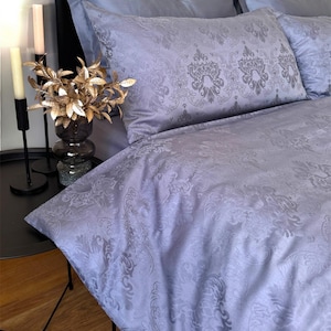May include: A light lavender bedding set featuring a tonal damask pattern on the duvet cover and pillowcases. The fabric has a subtle sheen, and the set is displayed on a bed with a black headboard, accompanied by two tall candles and a vase.