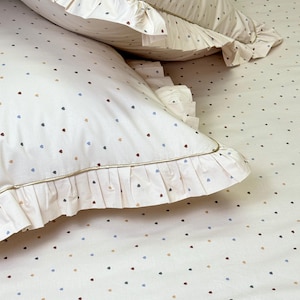 May include: Two off-white pillowcases and a matching bed sheet featuring a small, scattered heart pattern in muted shades of blue, brown, and tan. Each pillowcase is finished with a decorative ruffled edge and a thin, gold-toned piping.