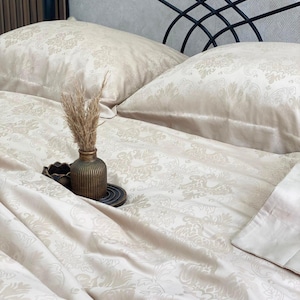 May include: A beige bedding set featuring a tonal damask pattern on the duvet cover and two pillows. A small, ribbed, dark brown vase containing dried grass sits on a dark, circular tray placed atop the rumpled fabric of the duvet cover.