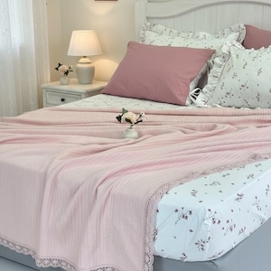 May include: A bed with a light pink blanket and matching pillow. The blanket has a lace trim. The sheets have a floral pattern. A white nightstand with a lamp and flowers is next to the bed. Decorative framed pictures are on the wall.