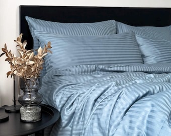 Puede incluir: Un juego de cama azul claro con un sutil patrón de rayas horizontales, que incluye una funda nórdica y fundas de almohada a juego. La tela tiene un acabado suave y ligeramente reflectante, creando una apariencia limpia y uniforme.