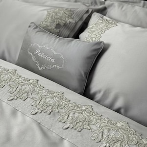 May include: A light gray bedding set featuring a rectangular throw pillow embroidered with the name Patricia in white script. The set includes matching pillowcases and a duvet cover, all decorated with intricate white floral lace trim.