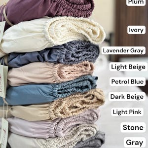 Puede incluir: Una pila de nueve sábanas bajeras de varios colores, etiquetadas como Plum, Ivory, Lavender Gray, Light Beige, Petrol Blue, Dark Beige, Light Pink, Stone y Gray. Cada sábana presenta un borde elástico y está doblada para mostrar la textura fruncida.