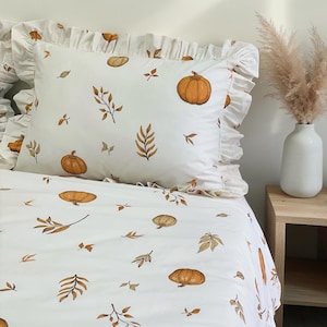 Puede incluir: Un juego de cama blanco con un patrón repetitivo de calabazas naranjas y hojas de otoño marrones. La funda de almohada tiene un borde con volantes y la funda nórdica muestra el mismo estampado botánico con varios tamaños de calabazas.